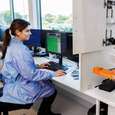 A female scientist in a blue lab coat sits at a computer beside a digital microscope scanning system.